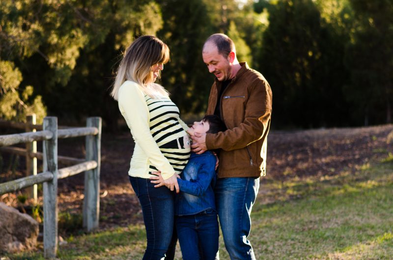 Imagen de una familia al atardecer en un parque verde de Murcia. Mamá está embarazada. Están de pie, mamá de perfil, muy pegada a ella su hijo, que le toca la barriga y la mira a la cara. Mamá lo mira a él también. Y papá está detrás de su hijo, acariciándole la cara y mirándole.